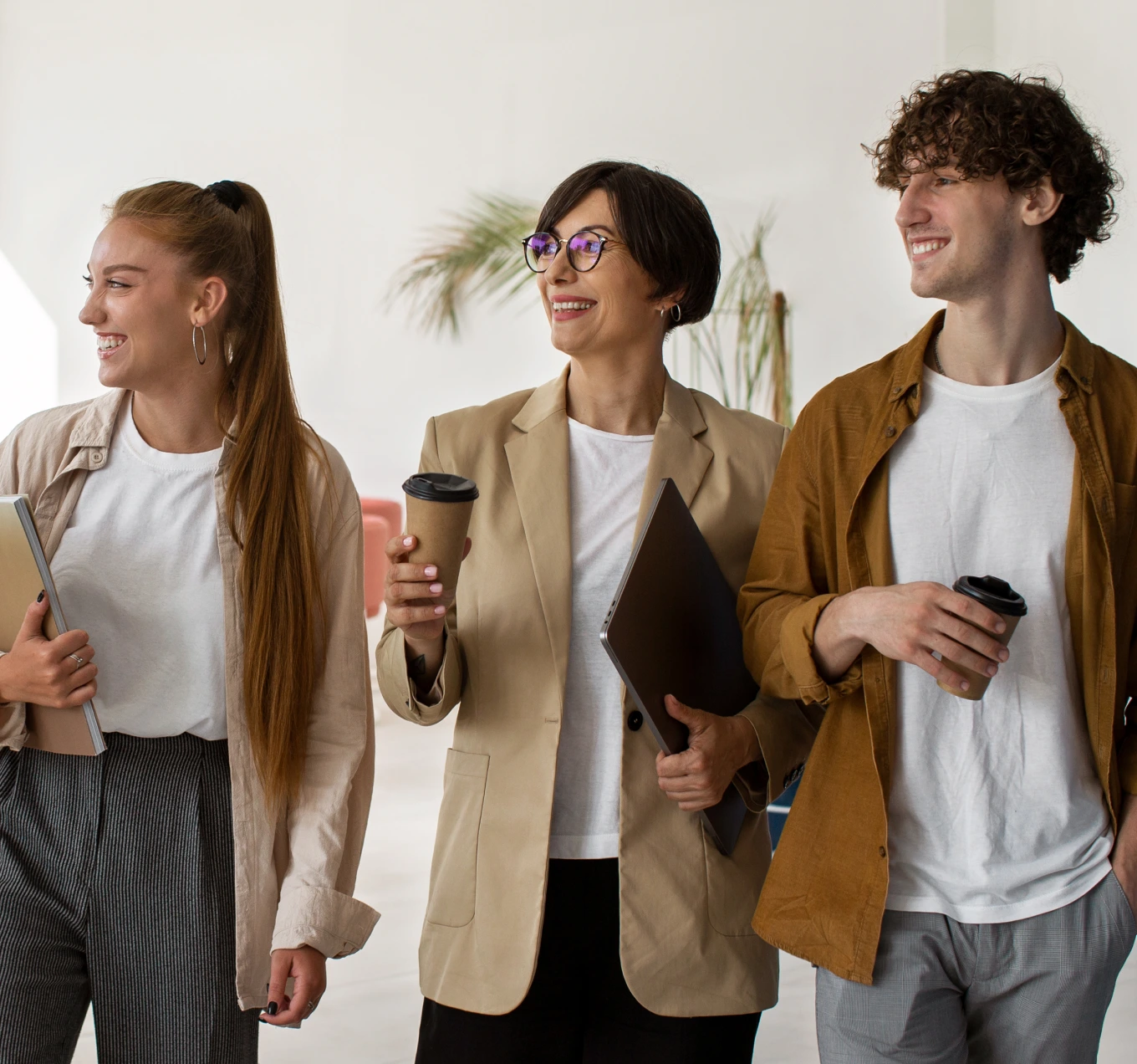 Three professionals standing indoors with coffee cups and a laptop, smiling