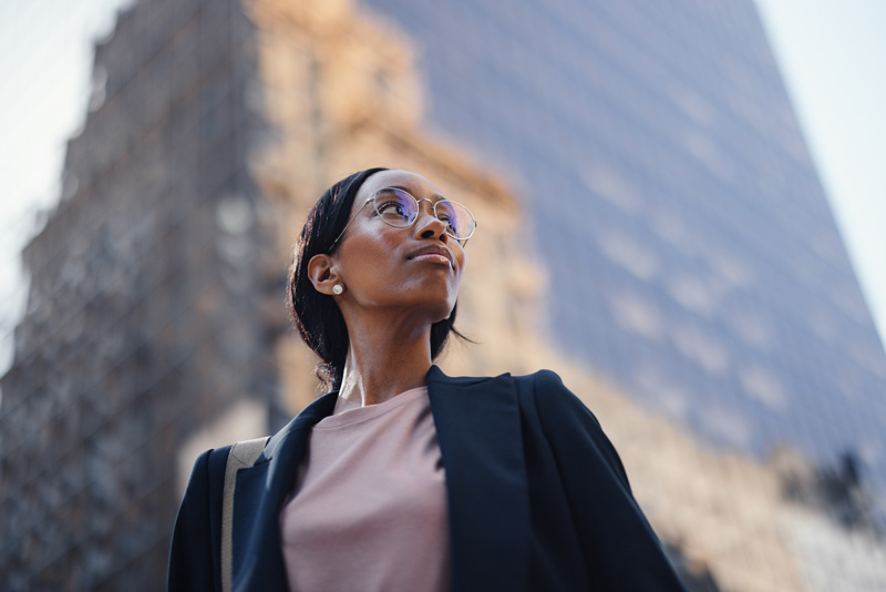 Confident woman wearing glasses and business attire looking upwards with a blurred tall glass building in the background.